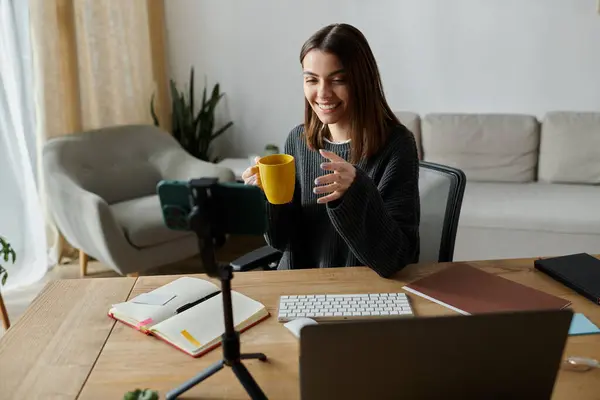 A young woman smiles as she films a video from her home office, enjoying a warm beverage. — Stock Photo