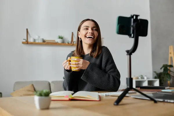 A young woman sits at a desk in her home, recording content on her phone, smiling happily and holding a cup. — Stock Photo