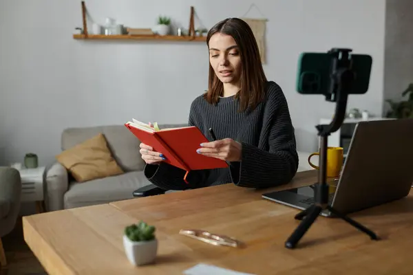 A young woman in a grey sweater sits at her desk, reviewing notes in a red notebook, while recording a video on her phone. — Stock Photo