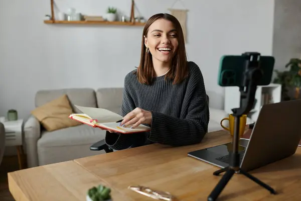 Uma jovem se senta em sua mesa, sorrindo e segurando um caderno, enquanto grava um vídeo para o público. — Stock Photo