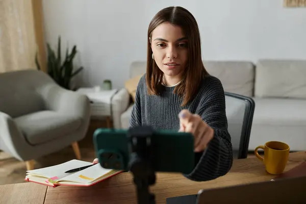 A young woman wearing a grey sweater sits at a desk in her home, filming a video on her smartphone. — Stock Photo