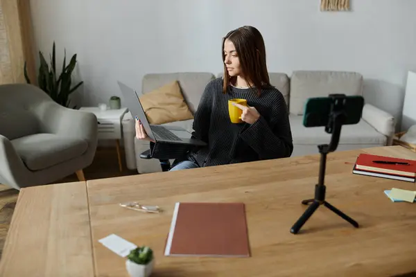 A young woman in a grey sweater sits at a table, working on a laptop and recording a video for her followers. — Stock Photo