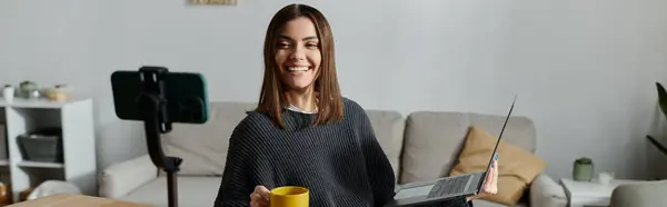 Una mujer joven se sienta en un sofá con una computadora portátil y una taza, sonriendo hacia un teléfono inteligente en un trípode. - foto de stock