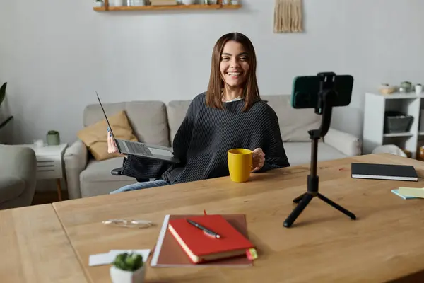 A young woman sits at a table with a laptop, a cup of coffee, and a phone on a tripod, creating content for her audience. — Stock Photo