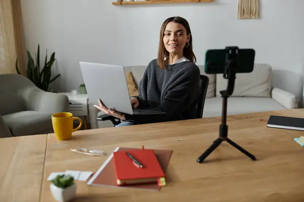 A young woman in a grey sweater works on her laptop at a desk in her home office, with a smartphone on a tripod in front of her. — Stock Photo