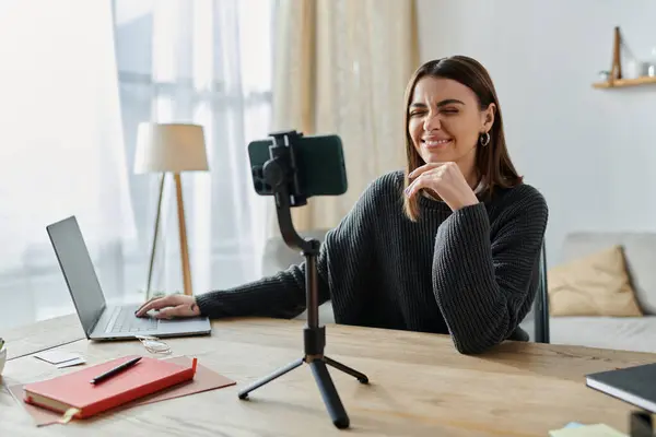 A young woman in a grey sweater records a video for her online audience while sitting at a desk with a laptop. — Stock Photo
