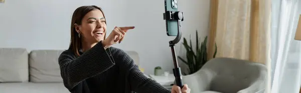 A young woman in a grey sweater sits on a couch and films a video with her smartphone. — Stock Photo