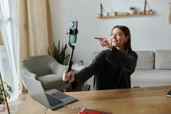 Una joven mujer se filma con un teléfono en un palo de selfie mientras está sentada en su escritorio de la oficina en casa. - foto de stock