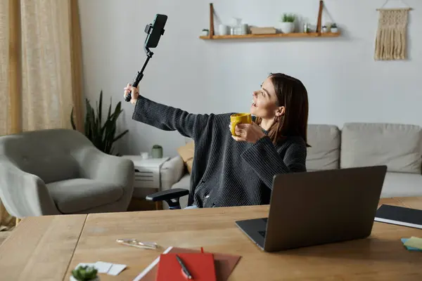 A young woman in a grey sweater films content with a selfie stick while sipping from a yellow mug. — Stock Photo