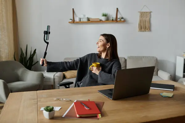 A young woman in a grey sweater smiles and holds a coffee mug while recording a video with her phone at a desk in a home office. — Stock Photo