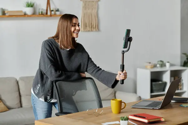 A young woman in a grey sweater films a video while sitting at a table in her home office. — Stock Photo