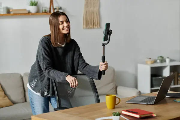 Молода жінка в сірому светрі фільми зі смартфоном на селфі палиці за стільцем за столом з ноутбуком. — стокове фото