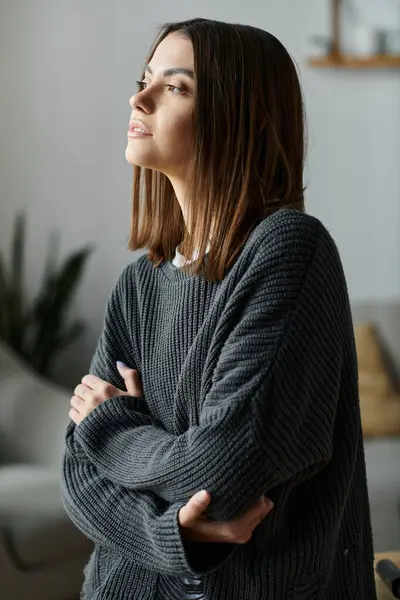 A young woman with brown hair, wearing a grey sweater, sits with her arms crossed, lost in thought while working from home. — Stock Photo