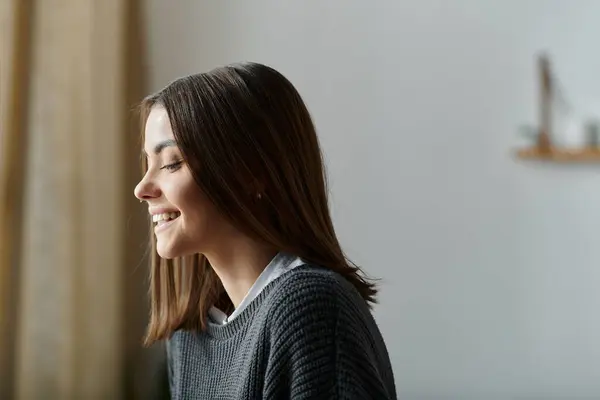 Uma jovem mulher em uma camisola cinza senta-se perto de uma janela, sorrindo e trabalhando em seu laptop. — Fotografia de Stock