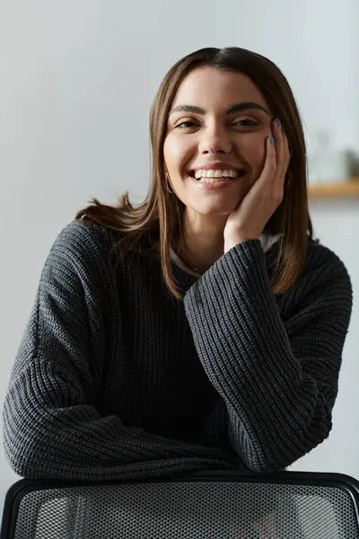 A young woman smiles brightly while working remotely from home, wearing a cozy grey sweater. — Stock Photo