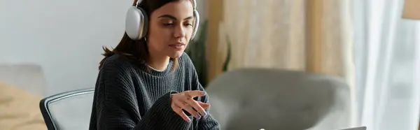 Uma jovem mulher usando fones de ouvido trabalha em um laptop em sua mesa de casa, com uma expressão focada. — Fotografia de Stock