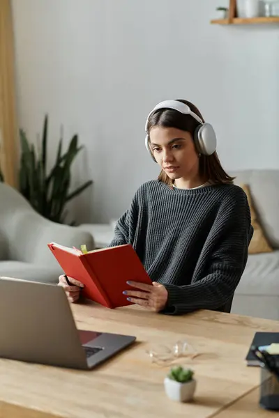 A young woman in a grey sweater works from home at a desk. She is wearing headphones and reading a red book while sitting at her desk. — Stock Photo