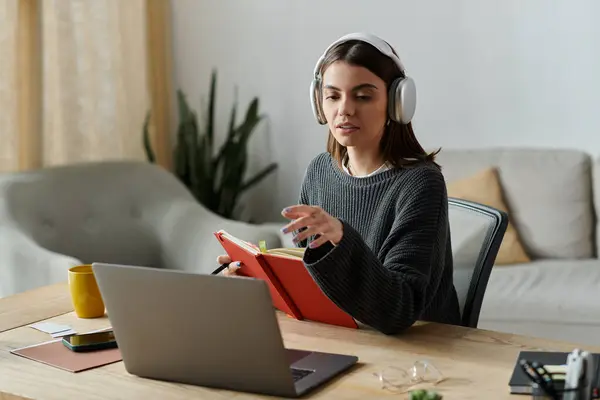 A woman wearing headphones works at her laptop, holding a red notebook open while looking at the screen. — Stock Photo