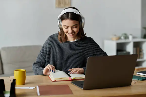 Une jeune femme portant un casque sourit alors qu'elle travaille sur son ordinateur portable et prend des notes dans un carnet. — Photo de stock