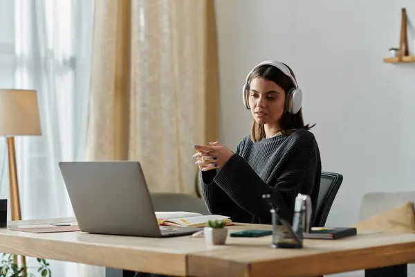 Uma jovem mulher de camisola cinza senta-se em uma mesa em seu escritório em casa, trabalhando em um laptop enquanto usa fones de ouvido. — Fotografia de Stock