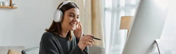 A young woman works remotely from home, wearing headphones and a grey sweater while creating content on her computer. — Stock Photo