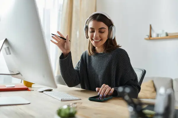 A young woman wearing headphones smiles and gestures while working at her desk in a home office. — Stock Photo