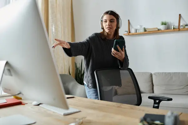 A young woman in a grey sweater stands in front of a computer, holding a phone, working from home. — Stock Photo
