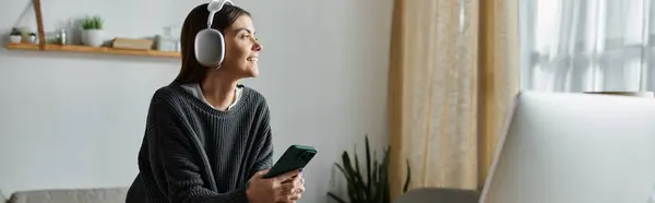 Una mujer sonriente en un suéter gris trabaja desde casa, usando auriculares y sosteniendo un teléfono inteligente. - foto de stock