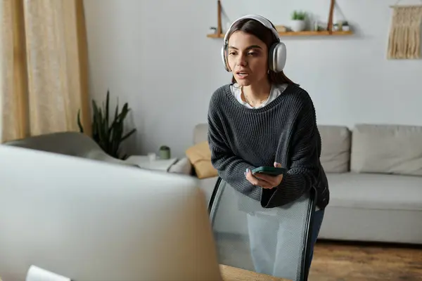 A young woman wearing headphones stands in a home office, looking at a phone and talking into a microphone. — Stock Photo