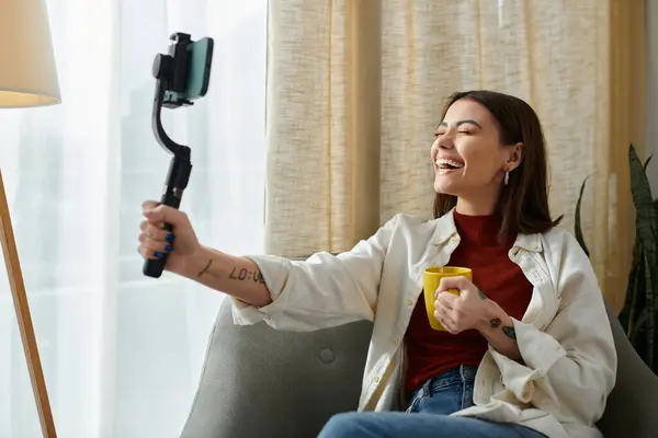 Cheerful influencer with tattoos and cup of coffee creating video content at home — Stock Photo