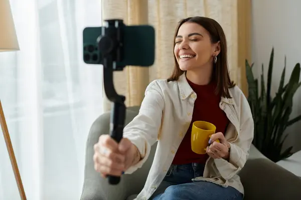 A young woman in casual attire sits on a couch, smiling as she vlogs while holding a smartphone stabilizer. — Stock Photo