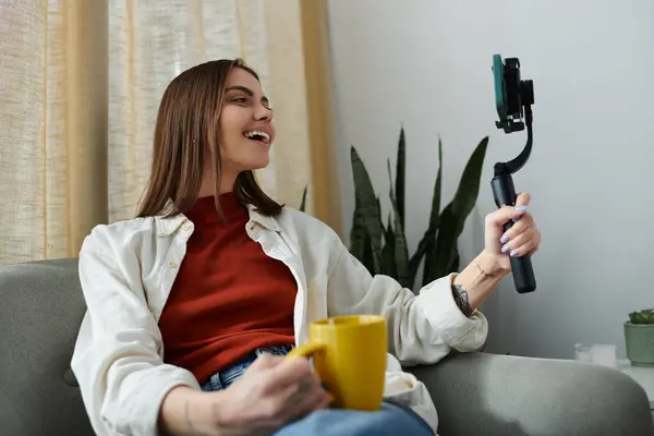 Uma mulher vestindo trajes casuais senta-se em um sofá, segurando um estabilizador de smartphones e filmando um vídeo enquanto sorri para a câmera. — Stock Photo