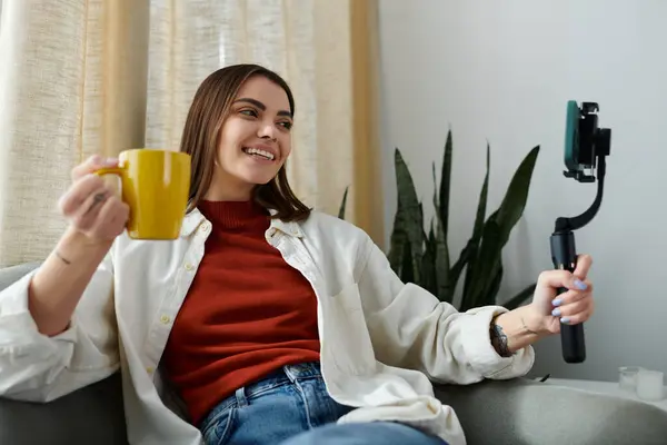 A young woman in casual attire sits on her couch and vlogs while holding a smartphone stabilizer. — Stock Photo