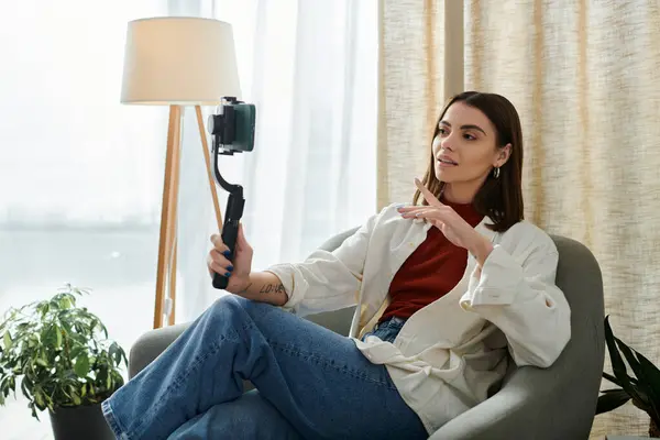 A young woman sits in a comfortable chair, holding a smartphone stabilizer and filming a vlog. — Stock Photo