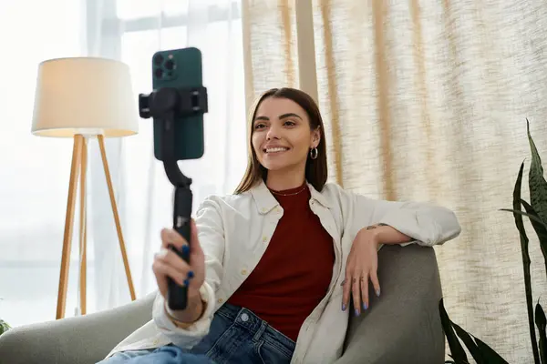 A young woman in casual attire films video content for social media while sitting on a chair in her home office. — Stock Photo