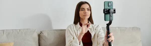 A young woman sits on a couch, holding a smartphone stabilizer and filming a vlog. — Stock Photo