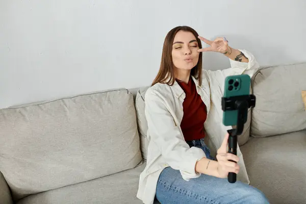 A young woman in casual attire sits on a couch and films a vlog with a smartphone stabilizer. — Stock Photo