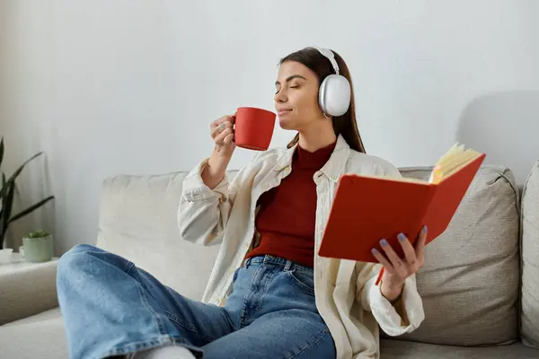 Una mujer joven con auriculares sorbe café mientras lee un libro en el sofá de su sala de estar. — Stock Photo