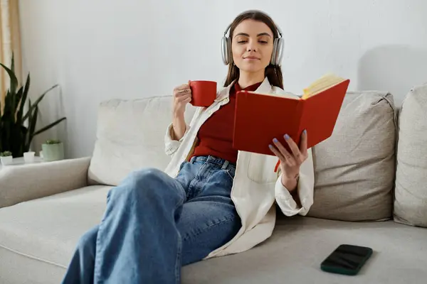 A young woman wearing headphones relaxes on a couch with a book and a cup of coffee. — Stock Photo