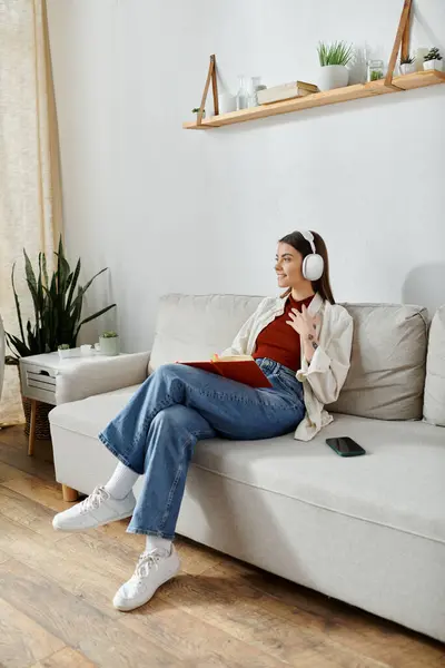 Una joven se relaja en un sofá blanco en una sala de estar, escuchando música con auriculares inalámbricos. - foto de stock