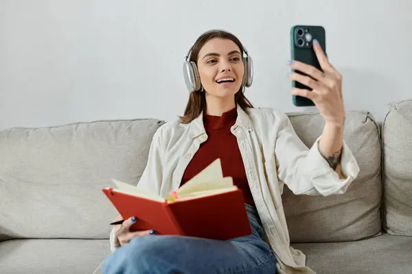 Молодая женщина сидит на диване в наушниках и держит книгу. Она улыбается, когда делает селфи со своим телефоном. — стоковое фото