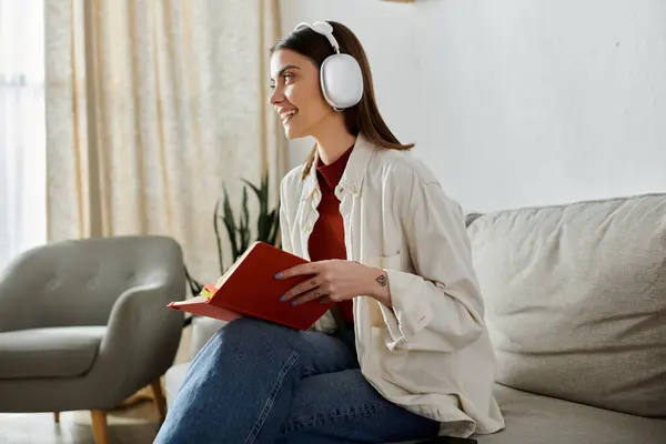 A young woman wearing headphones and a white shirt sits on a couch in a living room, holding a book and looking off to the side. — Stock Photo