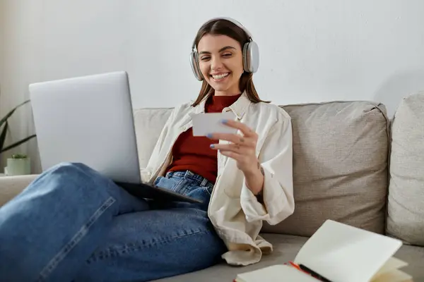 Uma mulher usando fones de ouvido senta-se em um sofá enquanto trabalha em seu laptop. Ela tem um cartão de visita na mão e sorri. — Fotografia de Stock