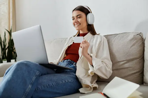 Una mujer joven, vestida con atuendo casual, se sienta en un sofá, trabajando en su computadora portátil mientras usa auriculares. - foto de stock