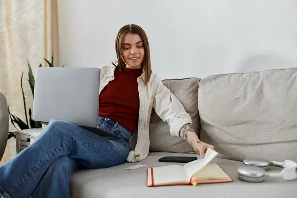 Une jeune femme en tenue décontractée travaille à distance sur son ordinateur portable depuis un canapé confortable à la maison. — Photo de stock