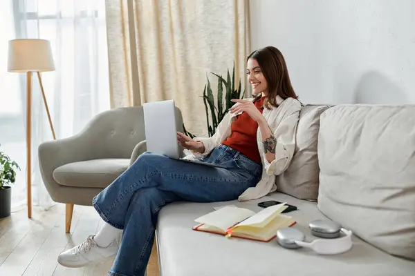 A young woman smiles as she works on her laptop from her living room couch, headphones nearby. — Stock Photo