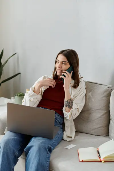 Una joven vestida de manera casual se sienta en un sofá, trabaja en su computadora portátil y habla por teléfono. - foto de stock