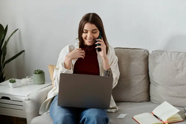A young woman sits on a couch, using a laptop and talking on a phone while working from home. — Stock Photo