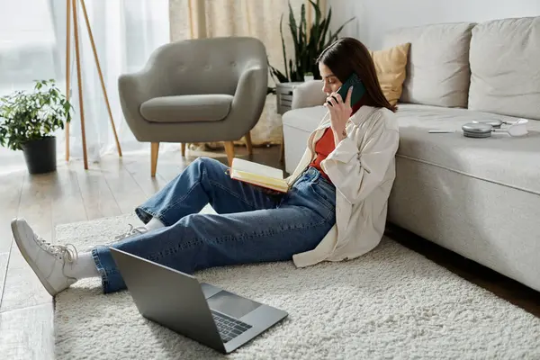 A woman sits on the floor in a casual outfit, talking on her phone while working on her laptop. — Stock Photo