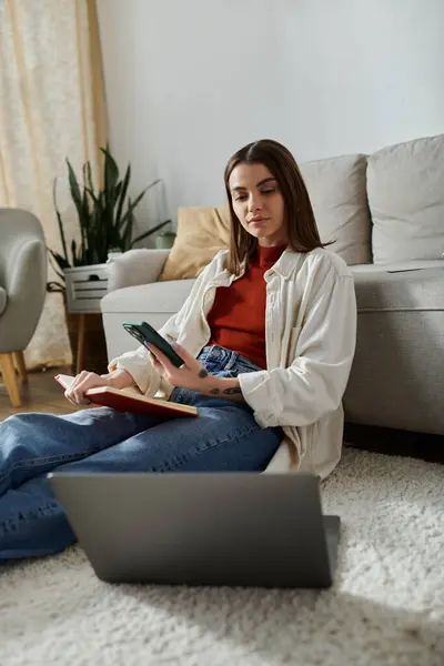 A young woman in casual attire sits on the floor with a laptop and phone in hand, working from home. — Stock Photo